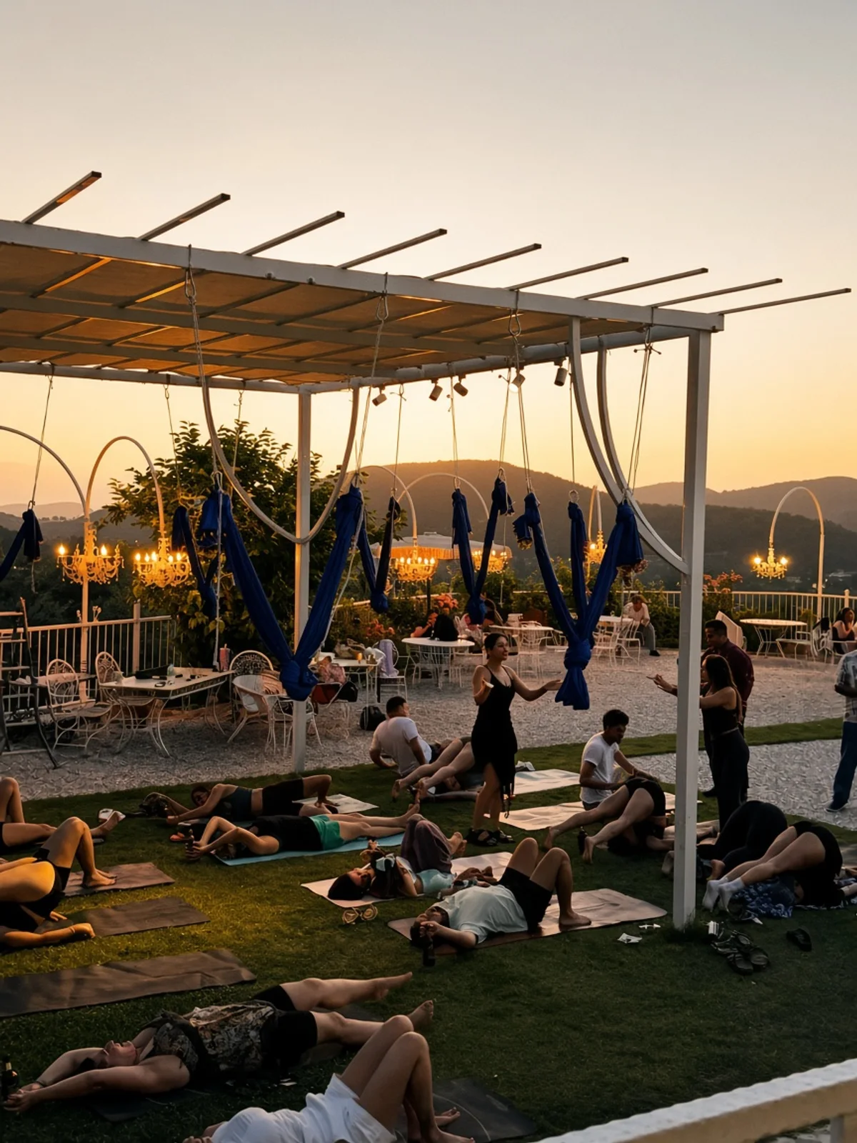 A group on mats under the pergola at sunset, blue silks overhead and olive hills beyond